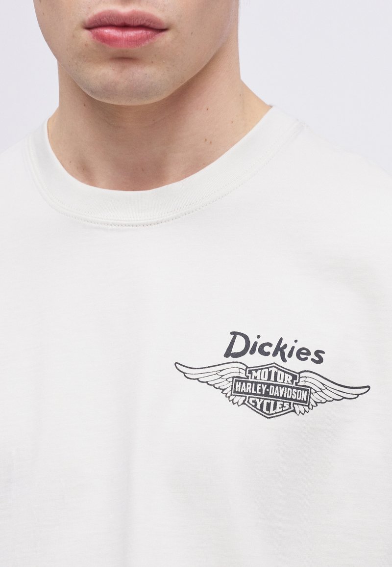 Gros plan d'une personne portant un t-shirt blanc avec le logo noir "Dickies" et le logo ailé de Harley-Davidson Motor Cycles sur la poitrine.