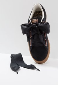 Baskets noires Puma avec des lacets en ruban satin noir épais noués en nœud, semelle gomme, et un lacet supplémentaire posé à côté sur une surface blanche.