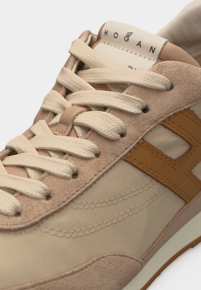 Gros plan d'une basket Hogan beige et fauve avec des lacets en tissu texturé, des panneaux en daim et des détails cousus sur le côté et la languette.