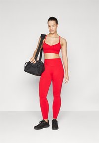 Rød sports-bh og matchende høytaljede tights laget av elastisk stoff, sammen med en svart treningsbag og svarte joggesko.