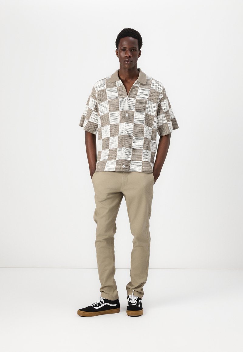 Short-sleeve button-up shirt features a checkered pattern in beige and white. Paired with beige pants and black sneakers with white accents.