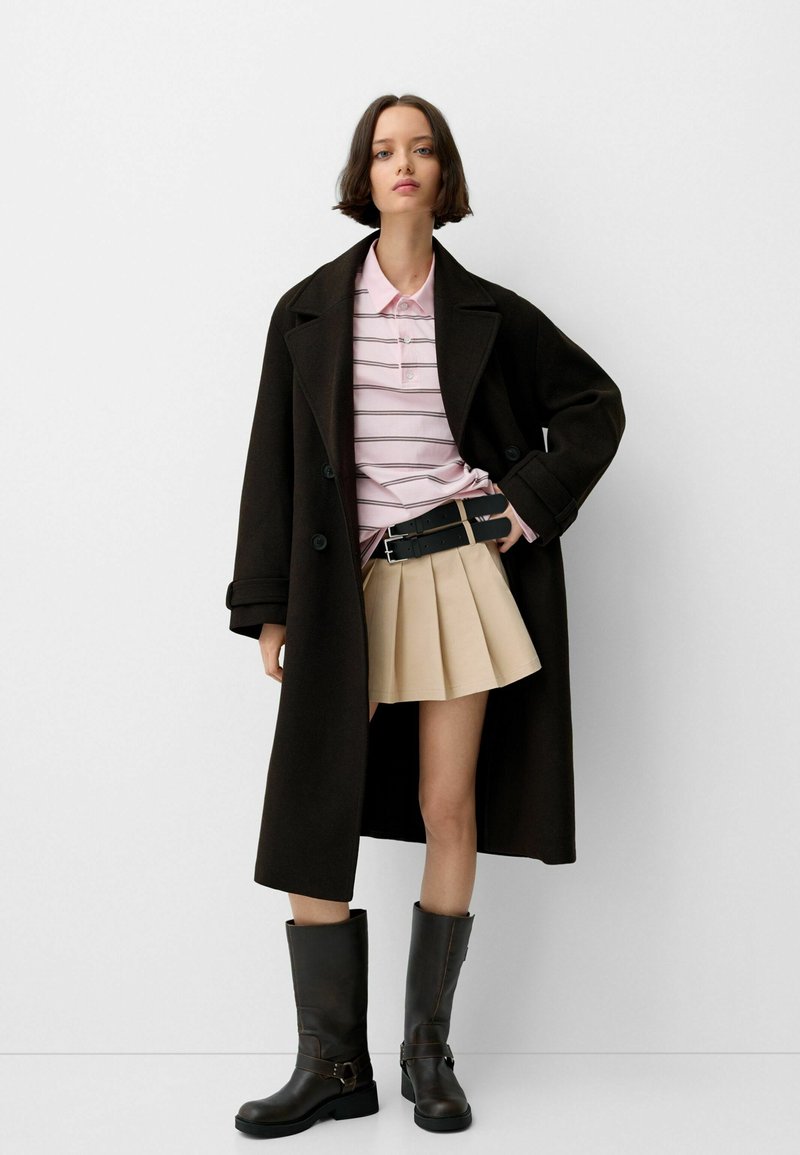 Czarny płaszcz, jasnoróżowa koszulka w paski, beżowa spódnica plisowana i brązowe kozaki za kolano z klamrami. Skupienie na czystych liniach i kontrastujących kolorach.