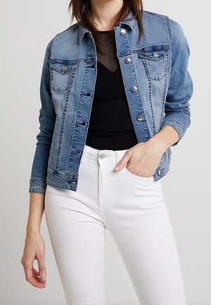 Femme portant une veste en denim bleue sur un haut noir et un pantalon blanc, une main dans la poche et l'autre détendue le long du corps.