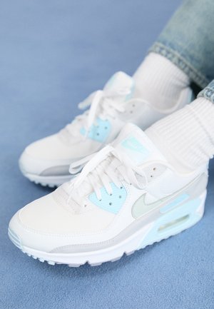 Witte en lichtblauwe Nike-sneakers gedragen met witte sokken en lichtblauwe jeans op een blauw tapijt.