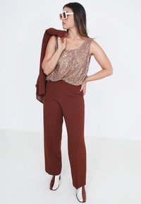 Gouden pailletten tanktop gecombineerd met bruine wijde broek. Accessoires zijn witte zonnebril en een bijpassende bruine jas.