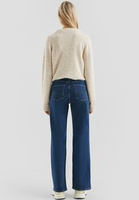 Lichtbeige textuursweater met een cropped achterkant, gecombineerd met blauwe wijde jeans. De outfit wordt voltooid met witte sneakers.