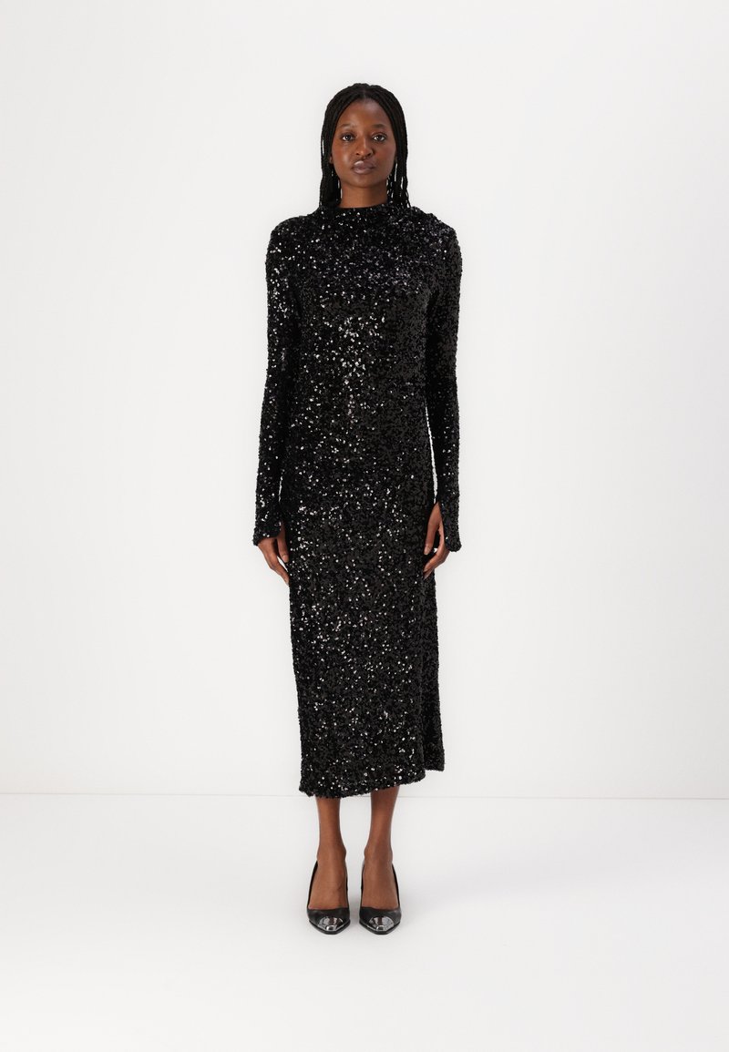 Robe noire à sequins, manches longues, col haut, silhouette ajustée, mi-longue, assortie à des talons noirs. Texture lisse avec des reflets brillants.