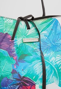 Sac fourre-tout avec des anses noires, imprimé de feuilles tropicales et de papillons dans des nuances de vert, bleu, violet, et une plaque de marque "Desigual" en argent.
