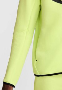 Camiseta de manga larga de un brillante color verde neón con una textura suave, con un ribete negro en los puños y el dobladillo, y un detalle de bolsillo lateral.
