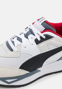 Sneaker bianco e grigio con accenti neri e mesh testurizzato. Presenta lacci bianchi piatti, colletto imbottito e un'etichetta del marchio ben visibile.