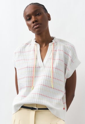 Frau trägt ein weißes, strukturiertes Kurzarmtop mit roten und gelben horizontalen Streifen und einem V-Ausschnitt mit Kordelzug, kombiniert mit beigen Hosen.