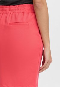Ołówkowa spódnica koralowa z elastycznym pasem i boczną kieszenią. Materiał wygląda na gładki i rozciągliwy, odpowiedni do codziennego lub swobodnego noszenia.