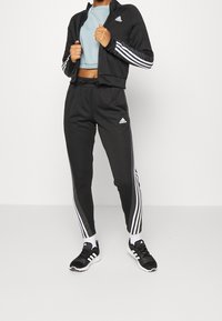 Schwarzer sportlicher Trainingsanzug mit weißen Drei-Streifen-Akzenten, Jacke mit durchgehendem Reißverschluss und schmal zulaufenden Hosen. Schwarze Sneaker runden den Look ab.