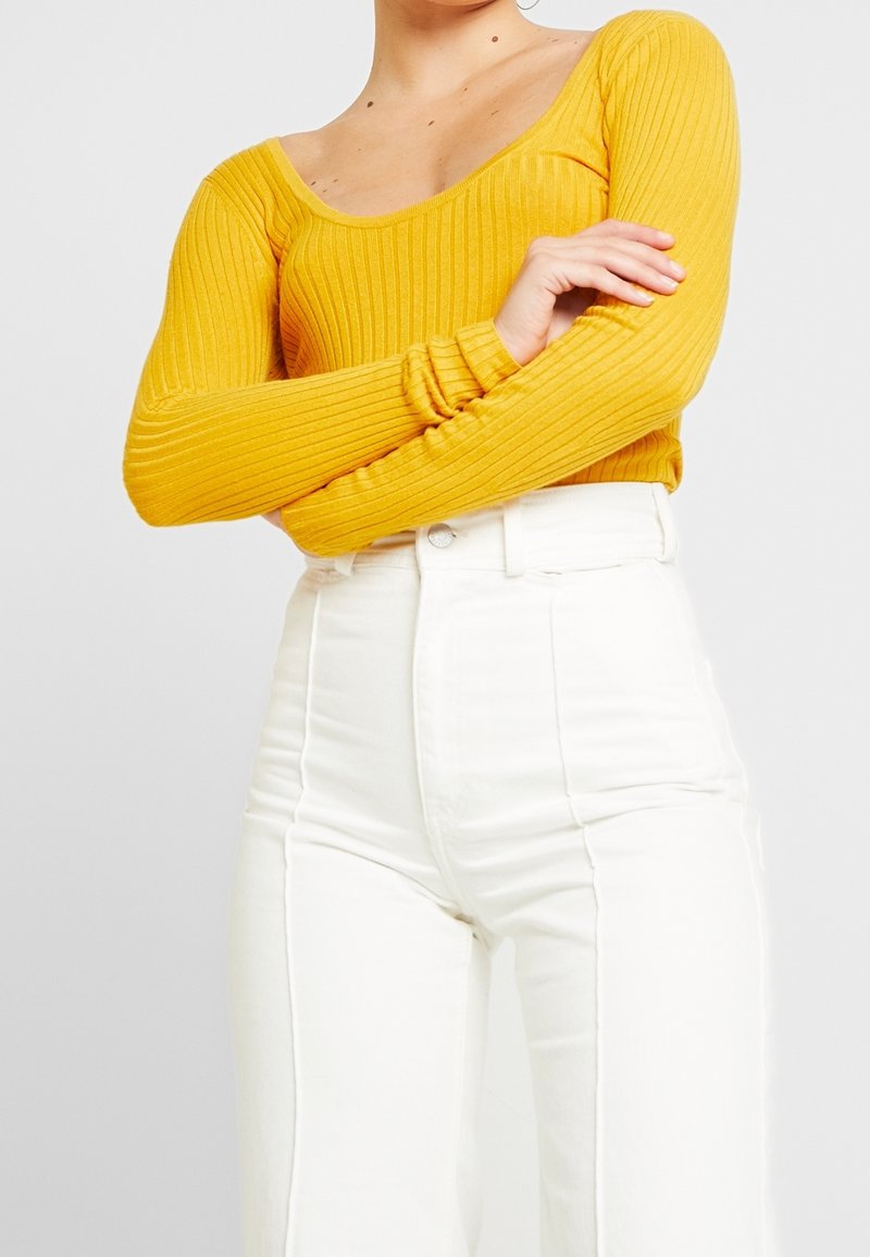 Persoon in een geribbelde mosterdgele lange mouw top en hoogwaisted witte broek met voornaad, met de armen over de borst gekruist.