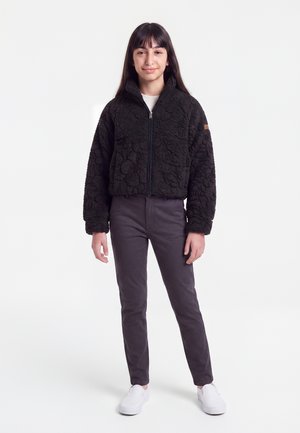 Adolescente aux longs cheveux foncés portant une veste zippée noire texturée, un pantalon foncé et des chaussures blanches sans lacets, debout sur un fond blanc.