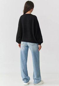 Czarny, oversizowy sweter wykonany z dzianiny z okrągłym dekoltem, w połączeniu z jasnoniebieskimi spodniami typu wide-leg i białymi butami. Tekstura swetra jest miękka.