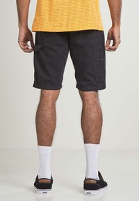 Black cargo shorts with side pockets, paired with black sneakers and white socks. A yellow patterned shirt is partially visible at the top.
