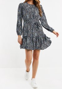 Vestido floral de manga larga con fondo negro, presenta patrones multicolores, un lazo en la cintura y un dobladillo con volantes en capas. Combinado con zapatillas blancas.