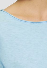 Haut bleu clair avec une texture douce et un grand col rond. Présente un ourlet cousu. Gros plan mettant l'accent sur le tissu et le col.