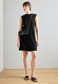 Fekete, magas nyakú, ujj nélküli ruhához textúrázott fekete clutch és fekete slide szandálok párosulnak. Világos fa padló háttér.