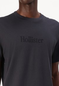 Juodas medvilninis marškinėliai su tekstūruotu, siuvinėtu "Hollister" logotipu priekyje. Apvali iškirptė ir trumpomis rankovėmis. Minkštas, laisvas stilius.