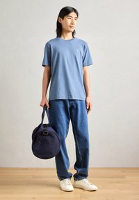 Modré bavlněné tričko, volný střih, kulatý výstřih; v kombinaci se světle modrými džíny. Drží kulatou námořnickou tašku. Bílé tenisky. Přírodní pozadí.
