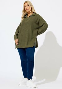 Übergroßer olivgrüner Strickpullover mit einem Viertel-Reißverschluss und seitlichen Schlitzen, kombiniert mit dunkelblauen Skinny Jeans und weißen Sneakers.