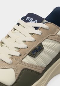 Κοντινό πλάνο μπεζ, λευκού και ελαιδί αθλητικού παπουτσιού FILA που εμφανίζει πλεκτή υφή, σουέτ επικαλύψεις, λευκά κορδόνια και λογότυπο της μάρκας στη γλώσσα και στο πλάι.