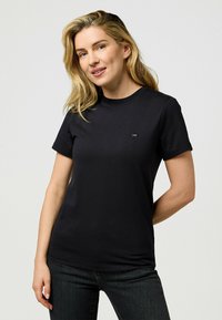 Sort bomulds t-shirt med korte ærmer, rund halsudskæring og et lille emblem på venstre bryst. Glat tekstur, tætsiddende pasform.