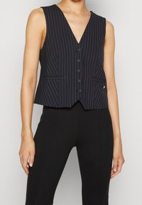Marinblå cropped väst med vertikala vita ränder, djup V-hals, sex knappförslutningar fram och två framfickor. Slät tygtextur.