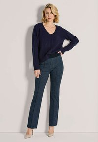Vrouw in een marineblauwe kabelgebreide trui, met gemusterde recht gesneden broek en beige hakken, staand met één hand op de heup tegen een effen achtergrond.