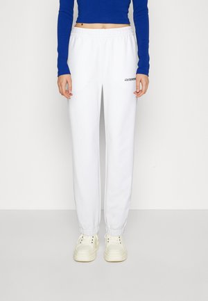 Persona che indossa pantaloni da ginnastica elasticizzati bianchi alla caviglia, top corto a maniche lunghe blu e sneakers platform panna, in piedi davanti a uno sfondo semplice.