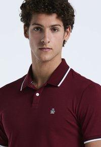 Bordeauxrotes Poloshirt aus Baumwolle mit einer Knopfleiste aus drei Knöpfen, weiß-blau gestreiften Kragen und einem kleinen Logo auf der Brust.