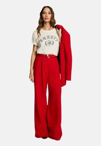 Pantalons larges rouges avec une taille ceinturée, associés à un t-shirt graphique blanc avec des lettres noires, complétés par un blazer rouge.