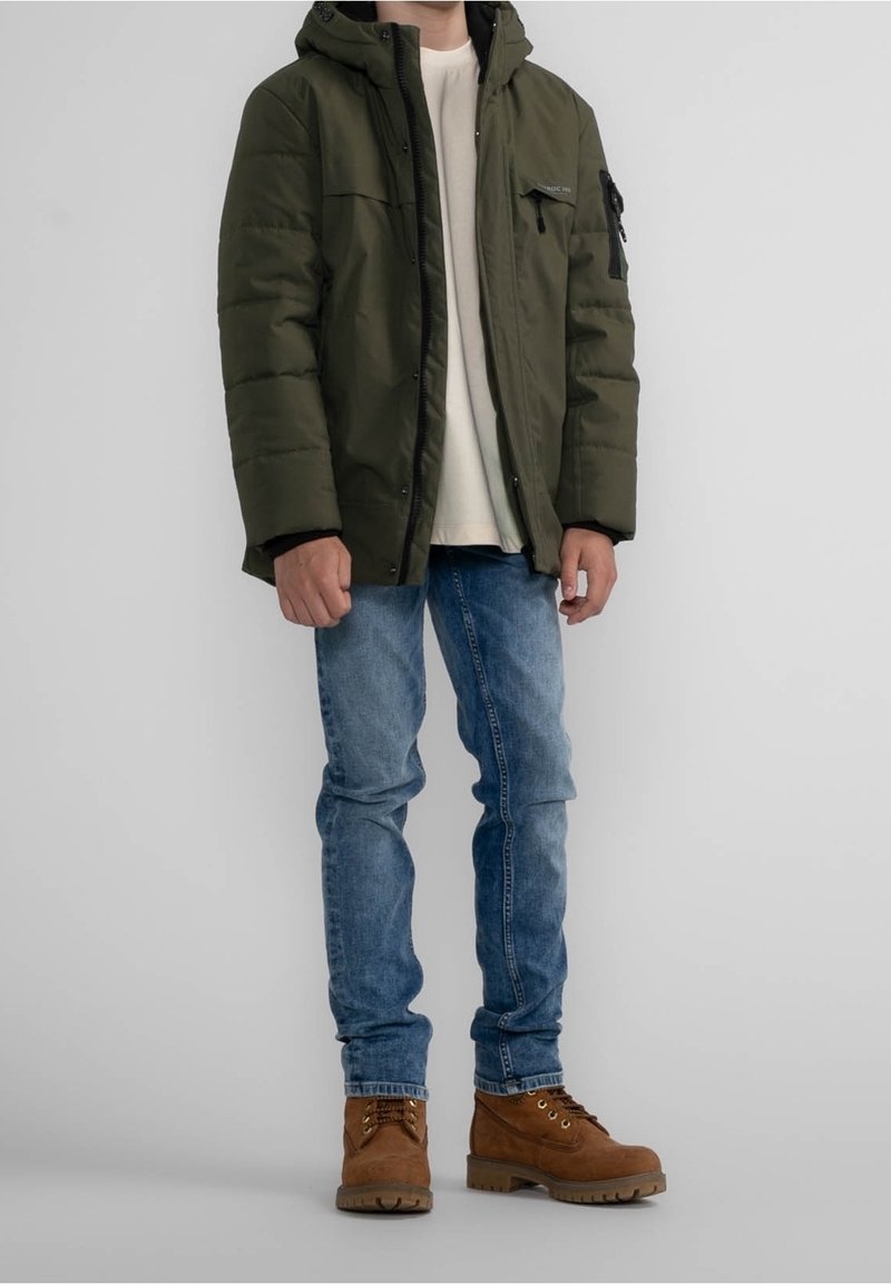 Personne portant une doudoune à capuche de couleur vert olive, une chemise beige clair, un jean bleu et des bottines marron à lacets, debout devant un fond gris.