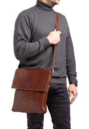 Bruine leren schoudertas met een klepdesign, een gladde textuur en een lange schouderband, gedragen over het lichaam door een persoon in een grijze trui.