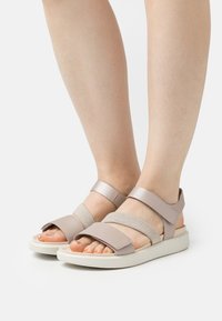 Lys beige sandaler med doble elastiske stropper og en flat hvit såle. Har et minimalistisk design og en åpen tå-struktur.