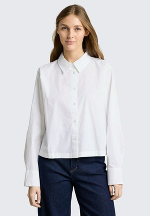 Junge Frau mit langen Haaren, die ein lockeres weißes Hemd mit Knöpfen und dunkelblaue Jeans trägt, vor einem schlichten hellen Hintergrund stehend.