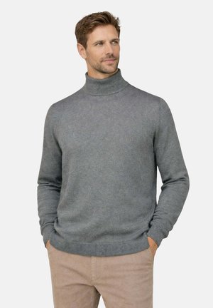 Uomo con maglione grigio a collo alto e pantaloni marrone chiaro, mani in tasca, che guarda leggermente di lato con sfondo neutro.