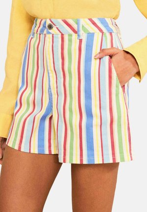 Kvinde iført shorts med høj talje med lodrette røde, blå, grønne, gule og hvide striber, kombineret med en gul bluse med lange ærmer.