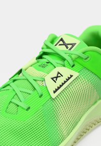 Neon zöld sportcipő, texturált hálós felsőrésszel, lapos fűzőkkel és jellegzetes geometriai díszítésekkel a nyelvén.