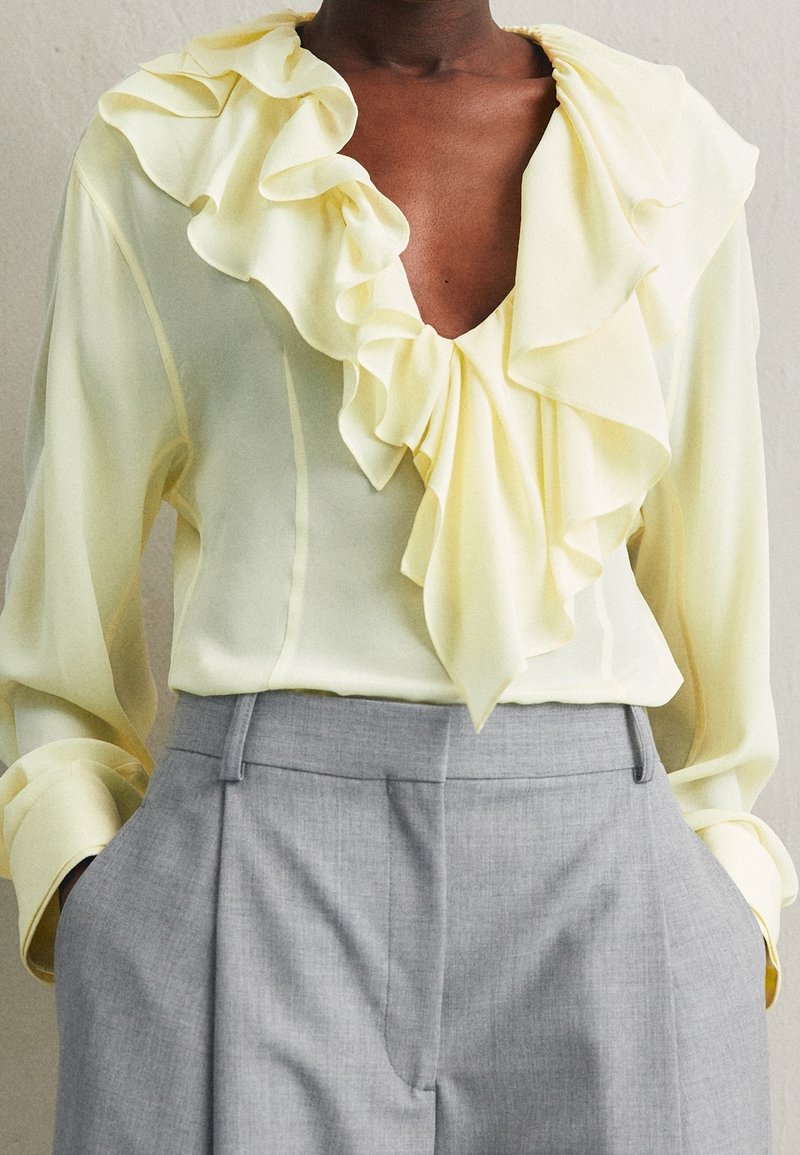 Person i en ljusgul blus med volangkrage och ljusgrå skräddarsydda byxor med händerna i fickorna.