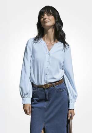 Mujer con los ojos cerrados, vestida con una blusa azul claro abotonada y una falda de mezclilla con un cinturón marrón, sosteniendo una bolsa en su mano izquierda.