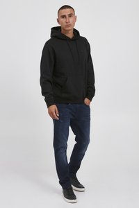 Felpa nera con cappuccio con tasca a marsupio e cordino, abbinata a jeans blu scuro. Le scarpe sono sneakers nere con suola bianca.