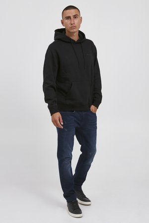 Zwarte hoodie met een buidelzak en capuchon met trekkoord, gecombineerd met donkerblauwe jeans. Schoeisel is zwarte sneakers met witte zolen.