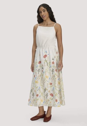 Frau mit langen dunklen Haaren, die ein weißes ärmelloses Kleid mit Blumenmustern und braune, gewebte flache Schuhe trägt, steht vor einem schlichten Hintergrund.