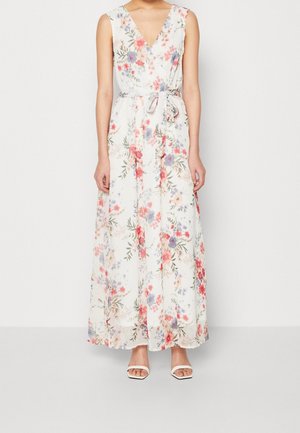 Femme portant une robe maxi blanche sans manches avec un motif floral coloré et une ceinture nouée à la taille, associée à des talons blancs ouverts devant.