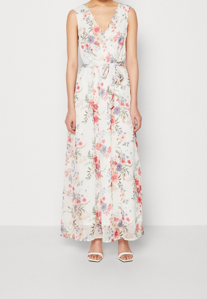 Femme portant une robe maxi blanche sans manches avec un motif floral coloré et une ceinture nouée à la taille, associée à des talons blancs ouverts devant.