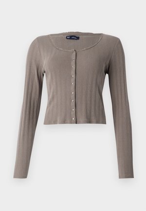 Μακριά μανίκια, κοντός καρυντινέ στην ανοιχτή καφέ ριμπ ύφανση. Διαθέτει στρογγυλή λαιμόκοψη και κούμπωμα με μεταλλικά στοιχεία.