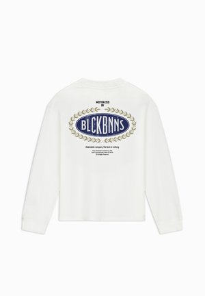Witte langarmshirt met een blauw ovaal logo, versierd met gouden laurieraccenten en tekst. Het kledingstuk heeft een ontspannen pasvorm en een ronde halslijn.